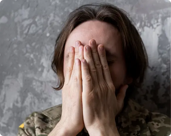 Person in camouflage military uniform covering face with hands against a textured gray wall.