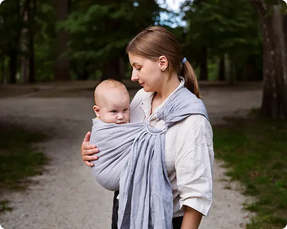Woman carrying a baby in a light gray ring sling outdoors on a gravel path with trees in the background.