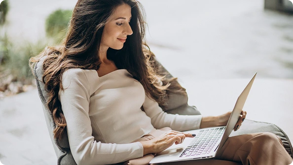 Woman with long brown hair sitting comfortably in a chair using a laptop outdoors.