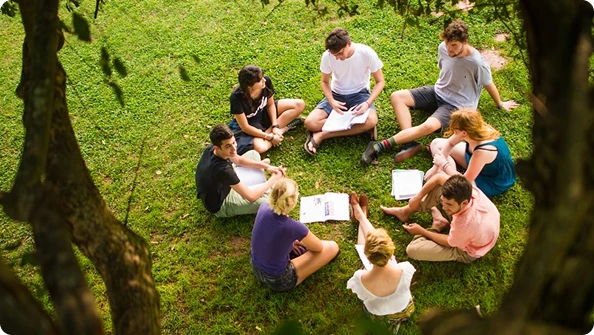 Group of seven young adults sitting in a circle on green grass outdoors, engaged in discussion with notebooks and papers.
