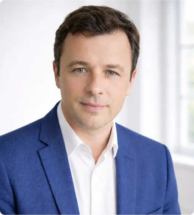 Portrait of a man with short brown hair and blue eyes wearing a blue blazer and white shirt, with a softly blurred background.
