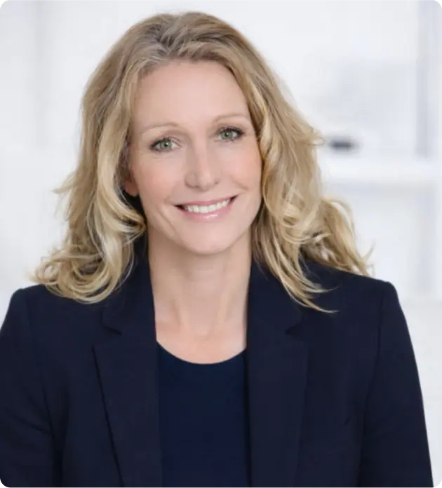 Smiling woman with blonde curly hair wearing a navy blazer and top against a light background.