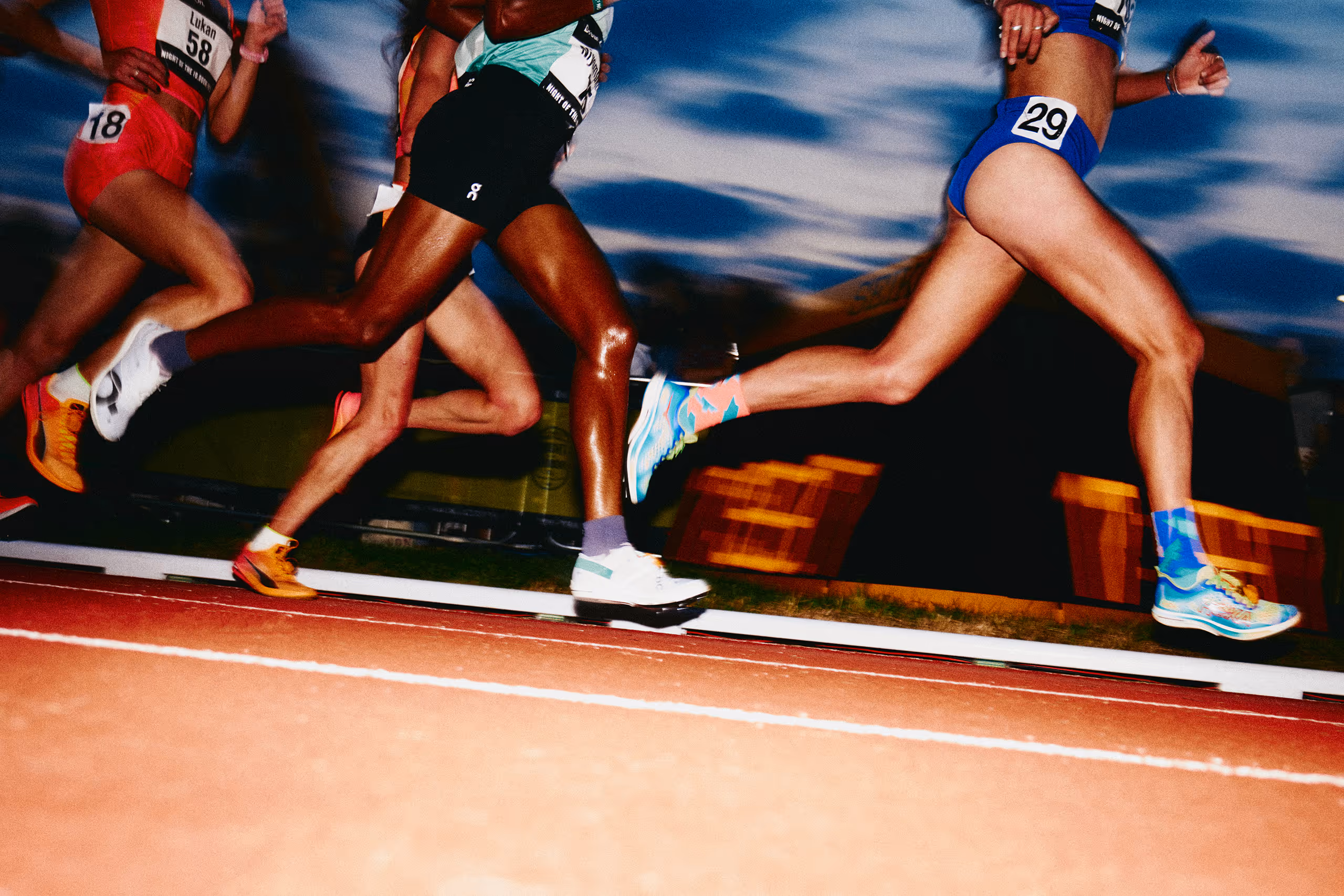 Close-up of runners’ legs on a track at night during a race, showing athletic footwear and race numbers.