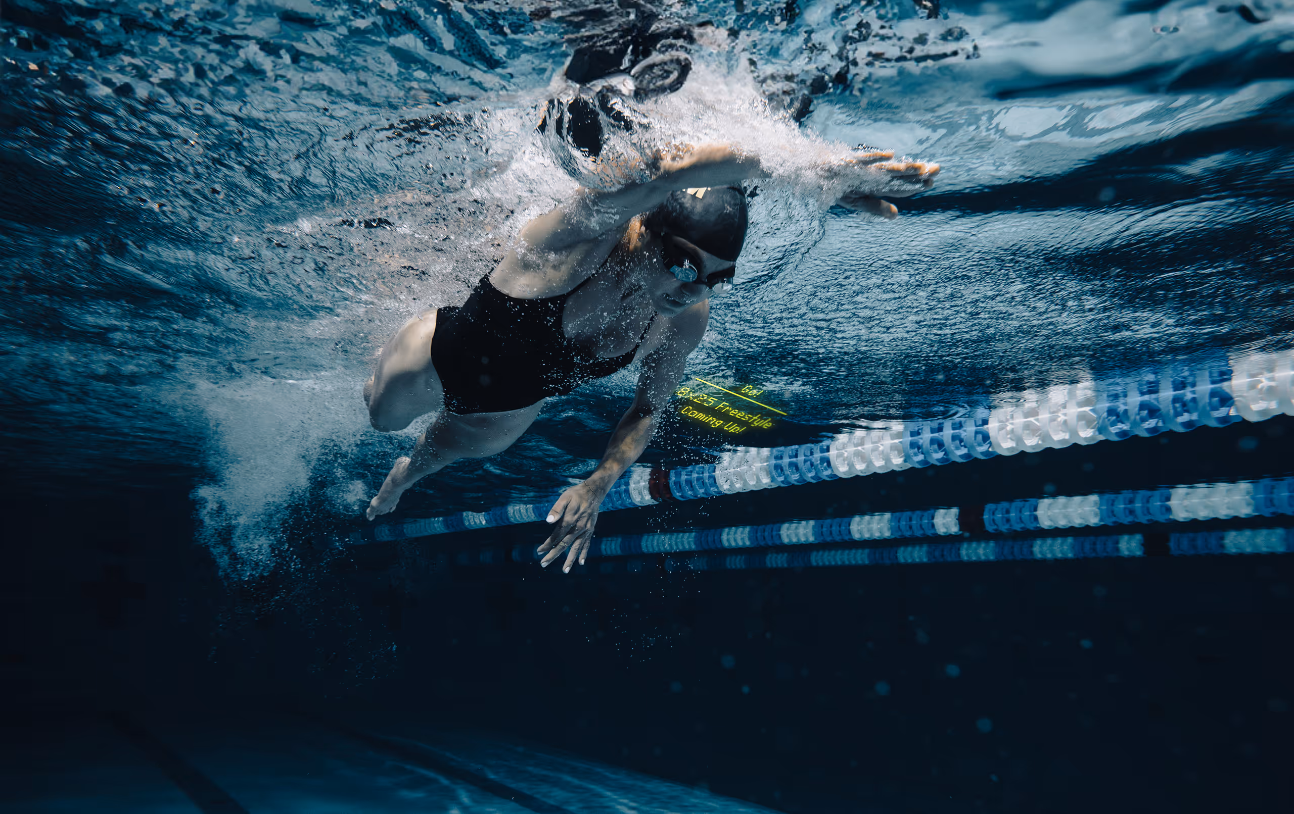 Female swimmer in a black swimsuit and goggles swimming freestyle underwater in a pool lane.