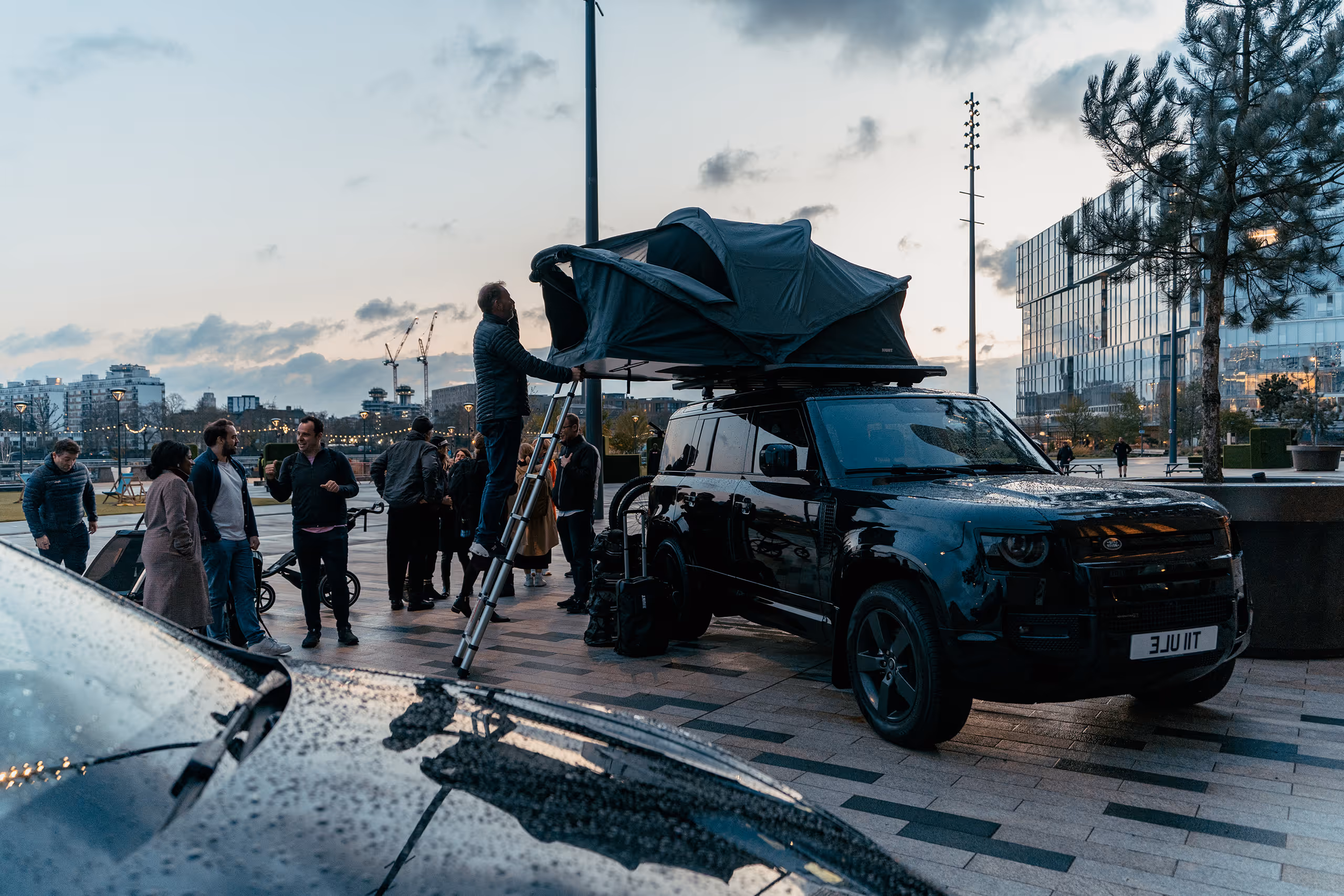Group of people gathered around a black SUV with a rooftop tent being set up using a ladder in a city plaza at dusk.