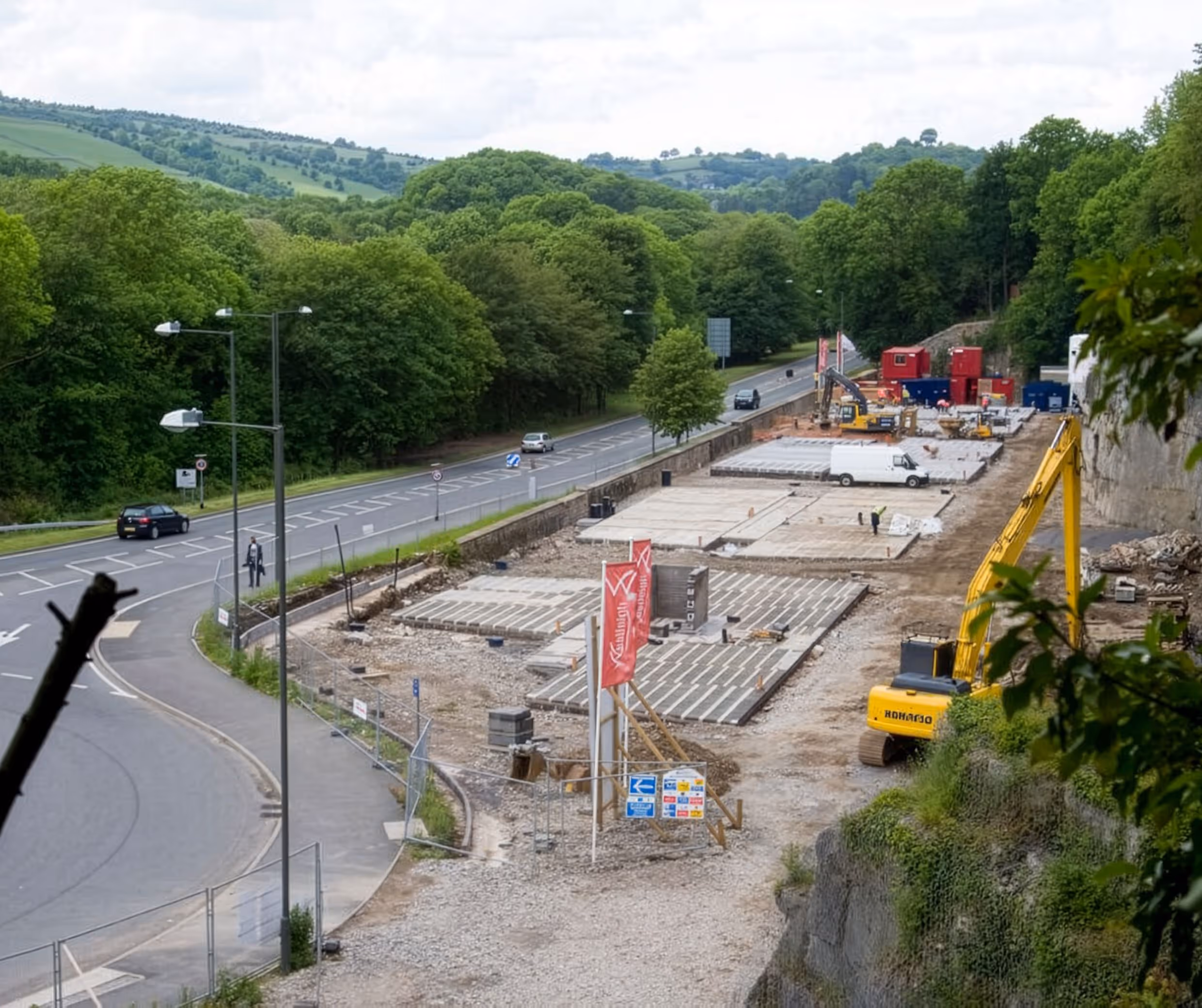 Construction site next to a road with green trees and hills in the background, featuring machinery and building materials.