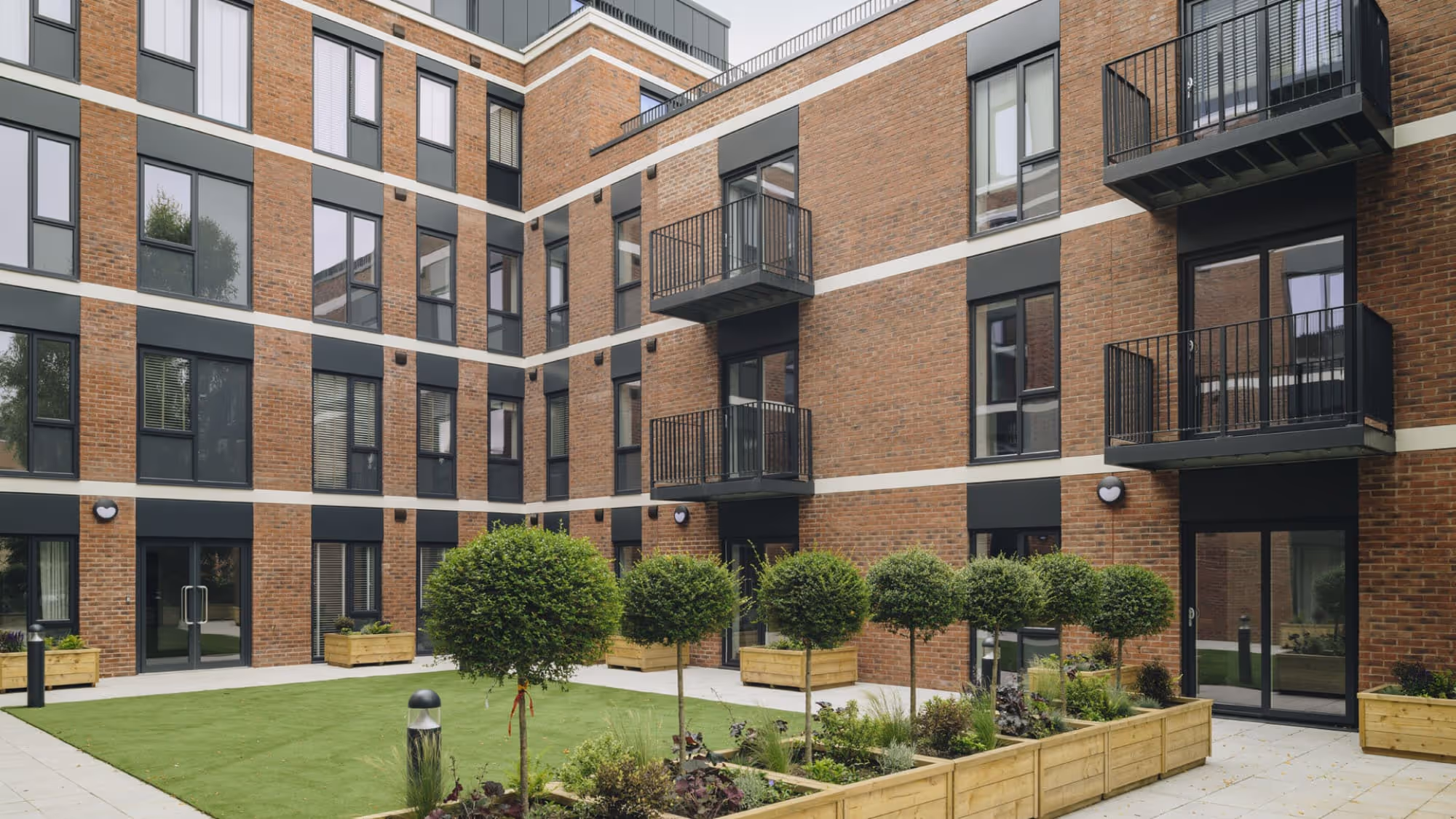 Modern brick apartment building with black-framed windows and balconies surrounding a courtyard with trimmed spherical shrubs and wooden planters.