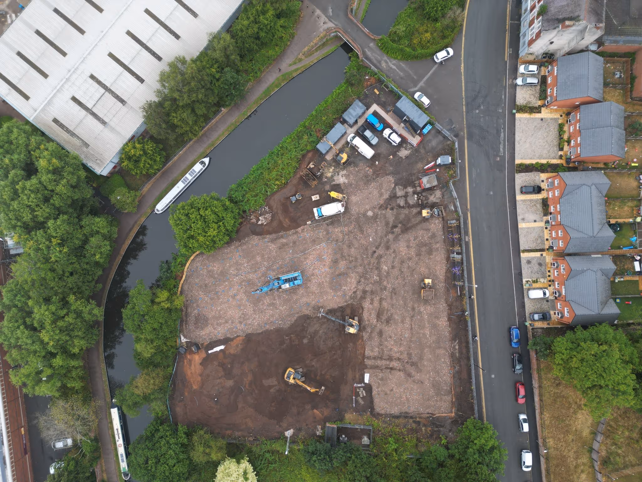 Aerial view of a construction site bordered by a canal with moored narrowboats on one side and residential houses with parked cars on the other.