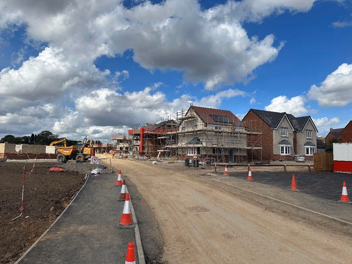 Residential development construction site with houses framed by scaffolding, construction vehicles, and a dirt road under a cloudy blue sky.