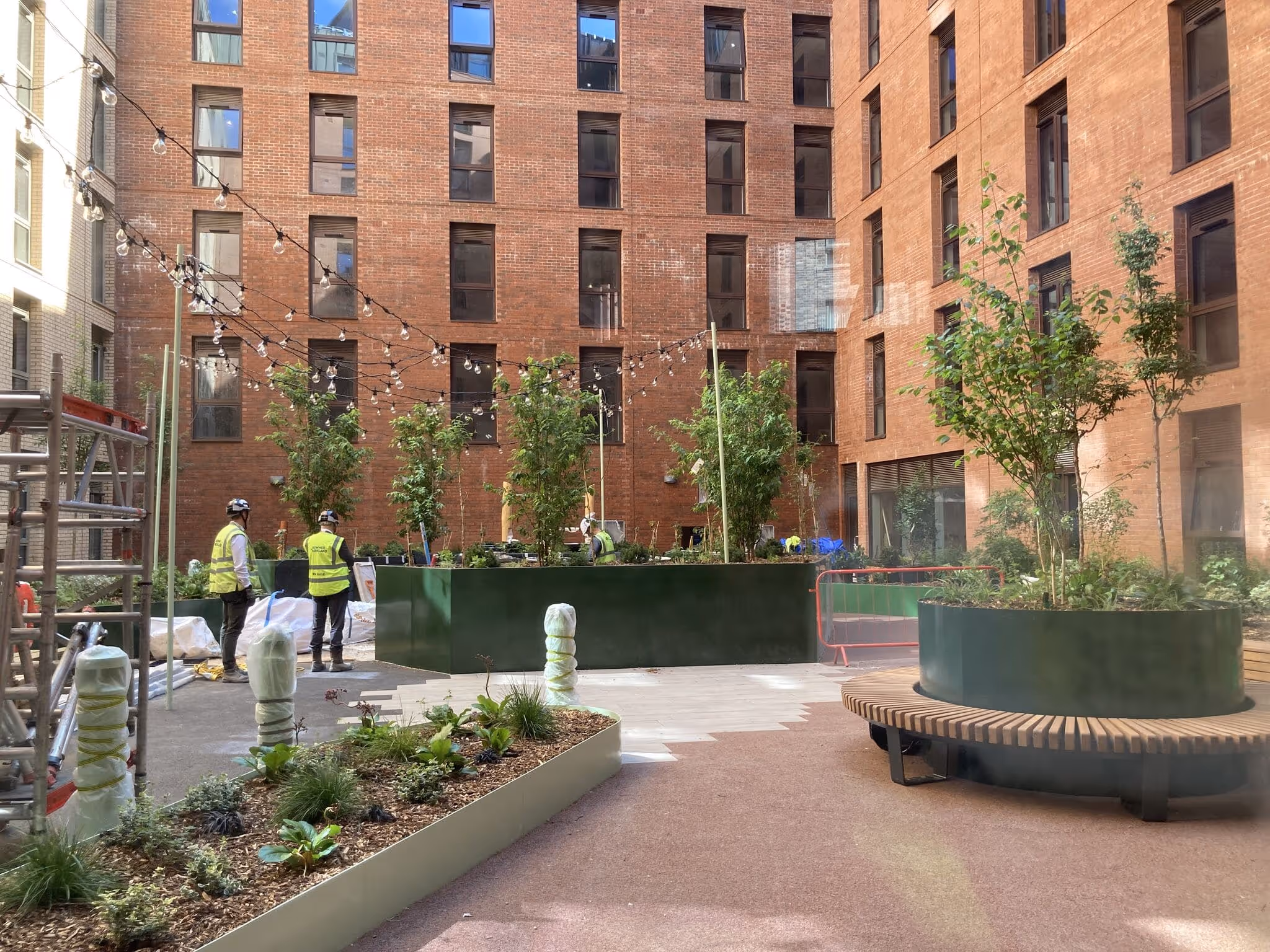 Courtyard garden with newly planted trees and plants in raised beds and circular benches, with workers in yellow safety vests and string lights overhead, surrounded by brick buildings.