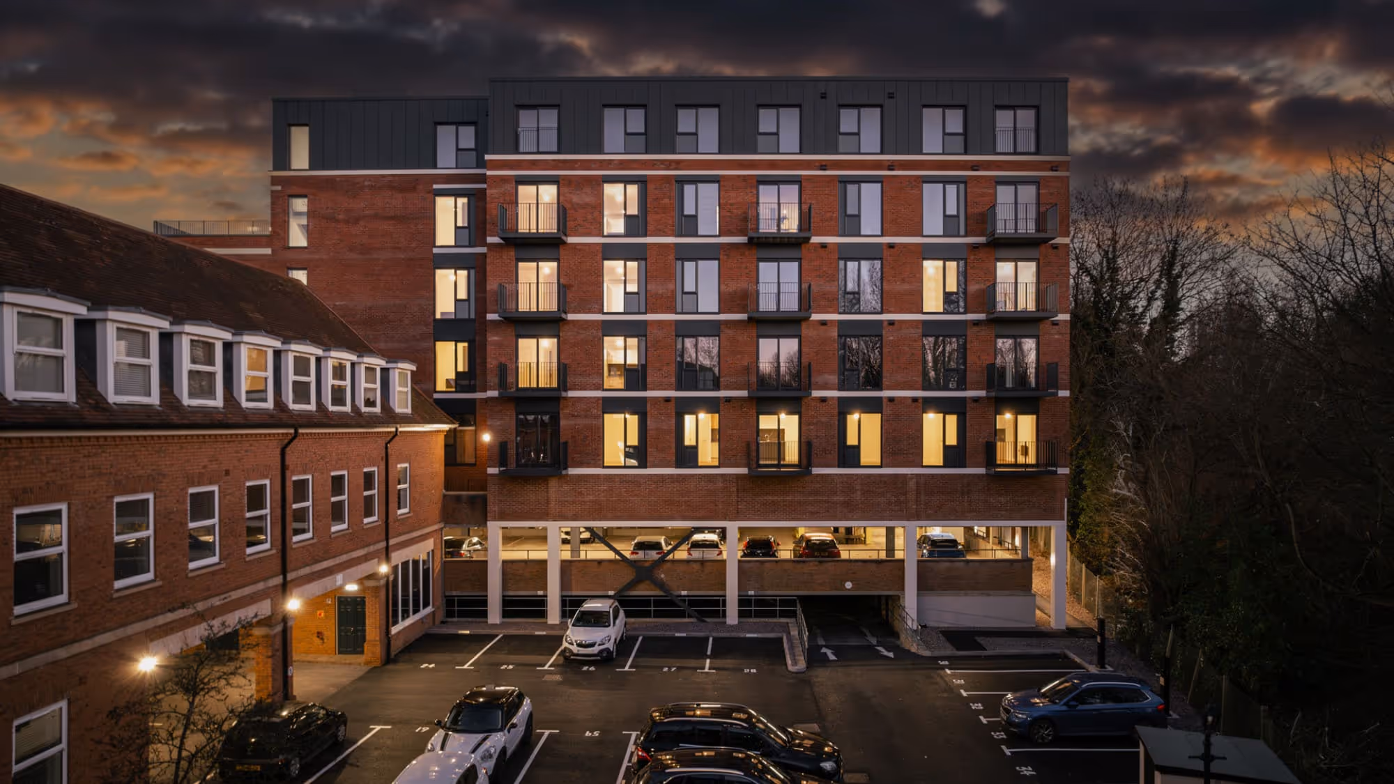 Multi-story brick residential building with lit windows and parked cars in front under a cloudy evening sky.