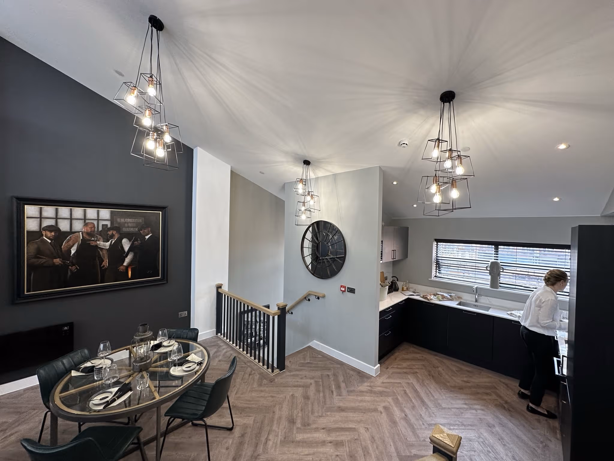 Modern dining and kitchen area with glass dining table set for four, contemporary pendant lights, large wall clock, and a person standing by the kitchen counter.