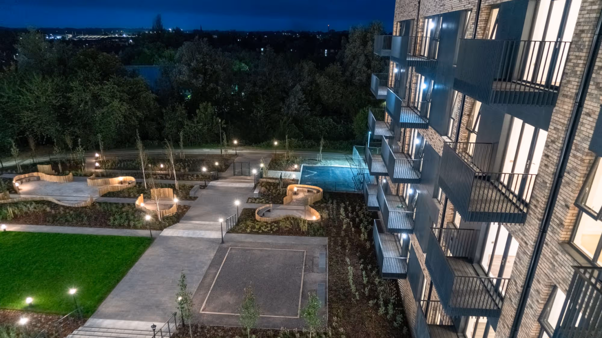 Night view of a modern apartment building with lit balconies overlooking a landscaped garden with curved wooden benches and pathways.