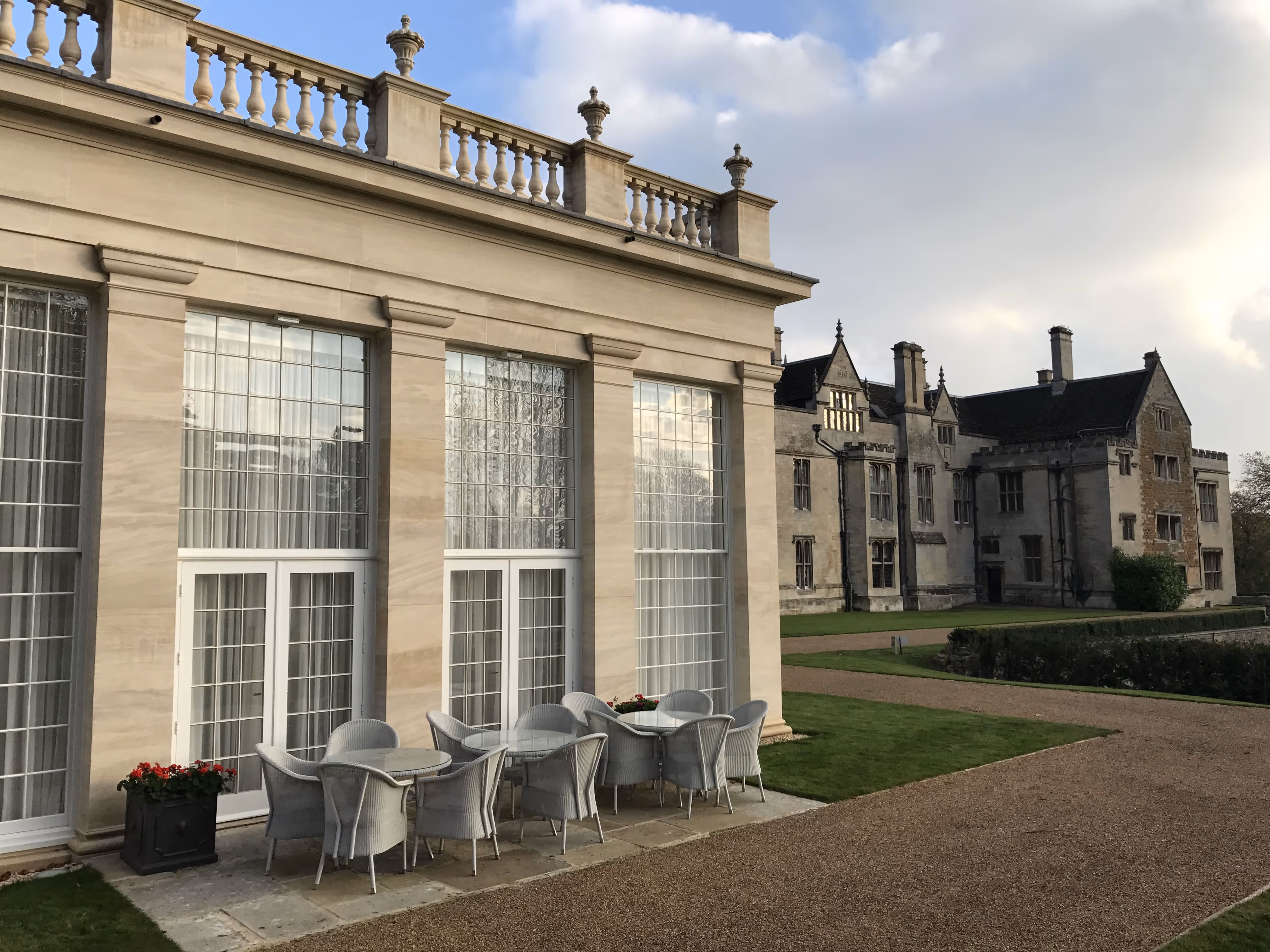 Outdoor patio with white tables and chairs beside large classical building with tall windows, next to a historic stone manor.