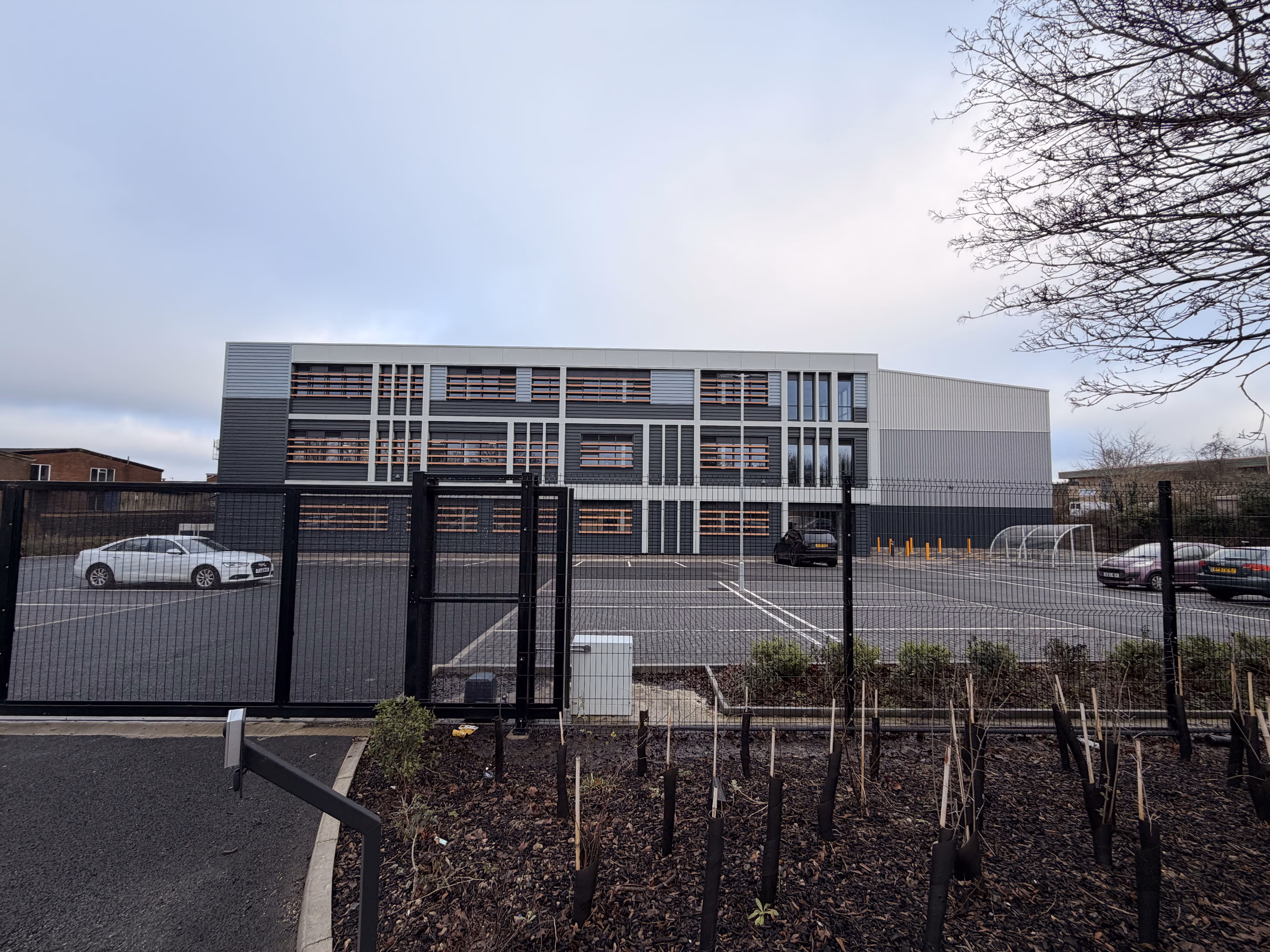 Modern three-story gray and white building behind a fenced parking lot with a few parked cars and young trees in the foreground.