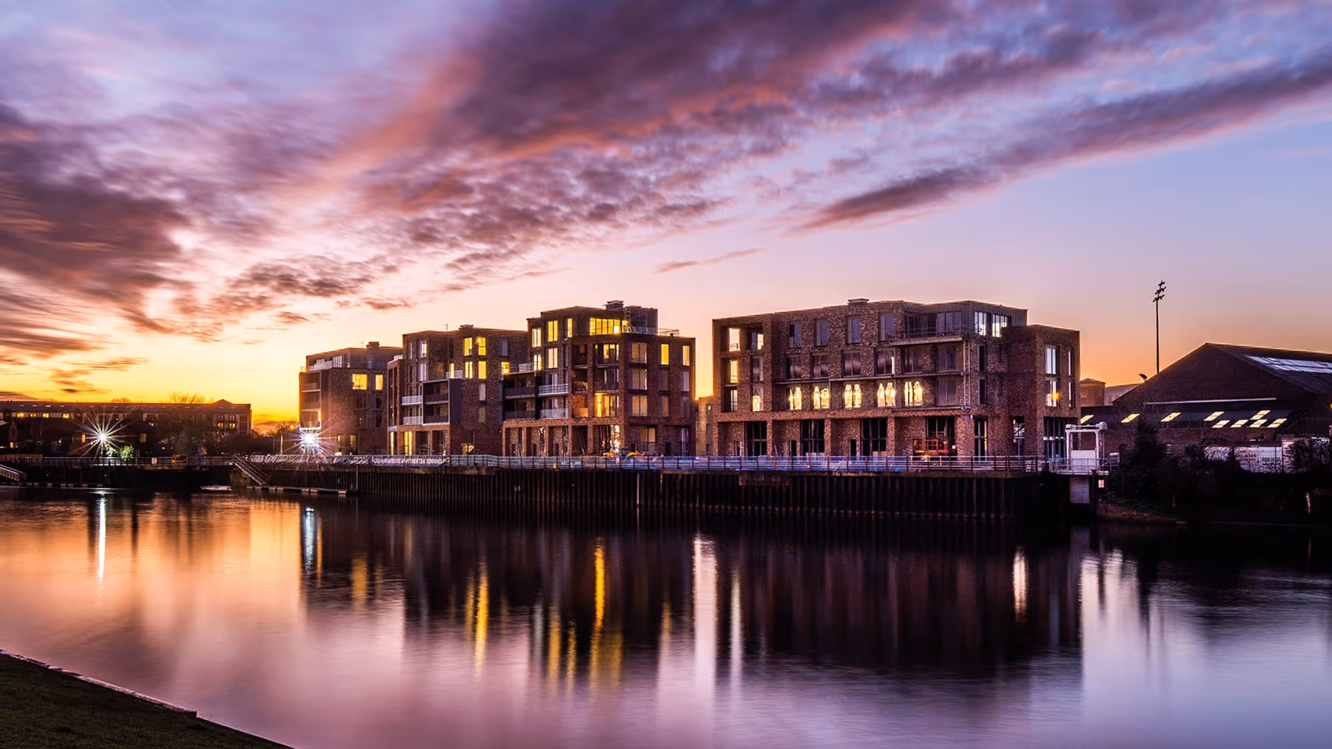 Apartment buildings by a calm river at sunset with colorful clouds and lights reflecting in the water.