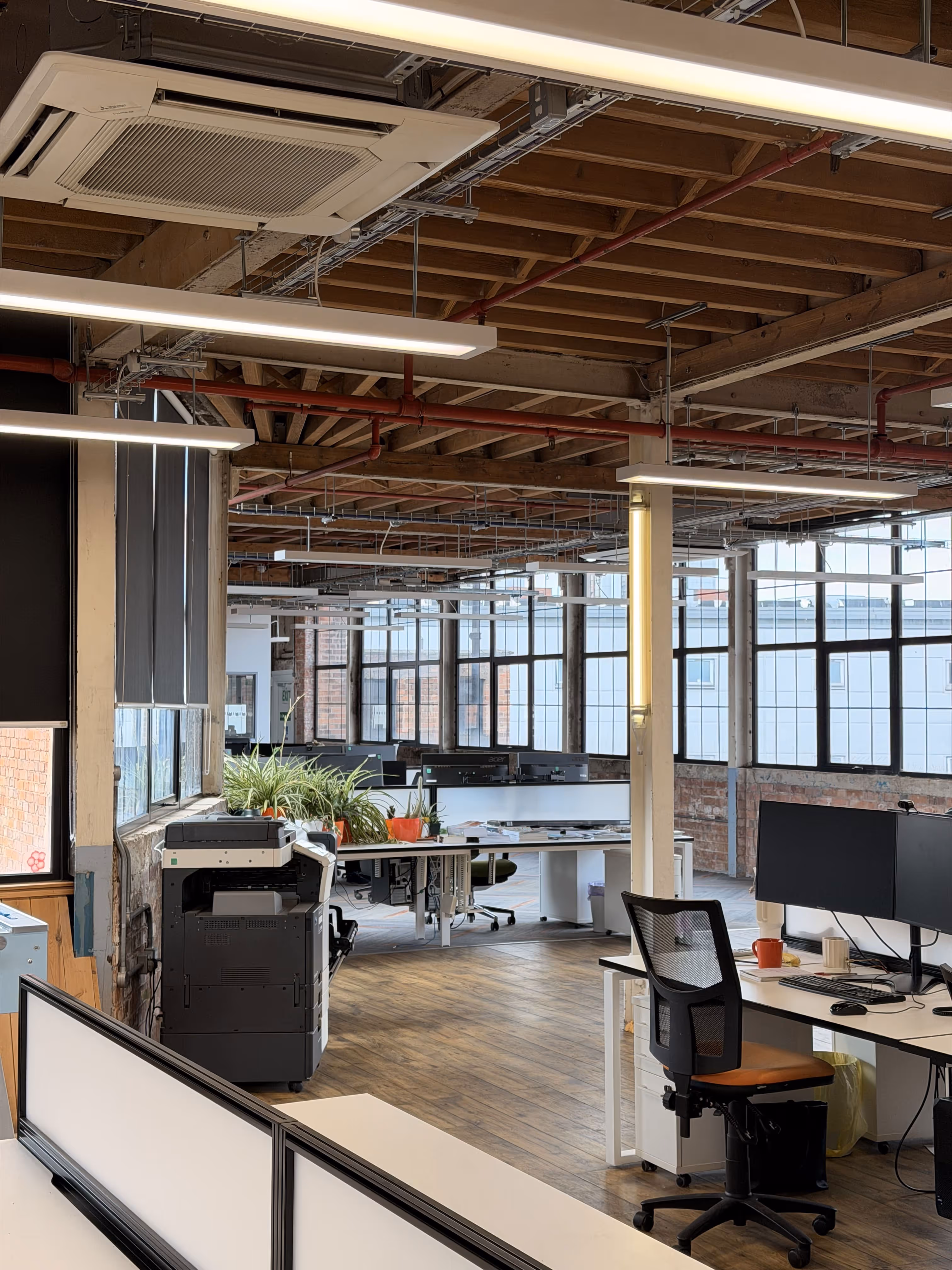 Modern open office with wooden ceiling beams, large windows, multiple desks, office chairs, plants, and a printer.
