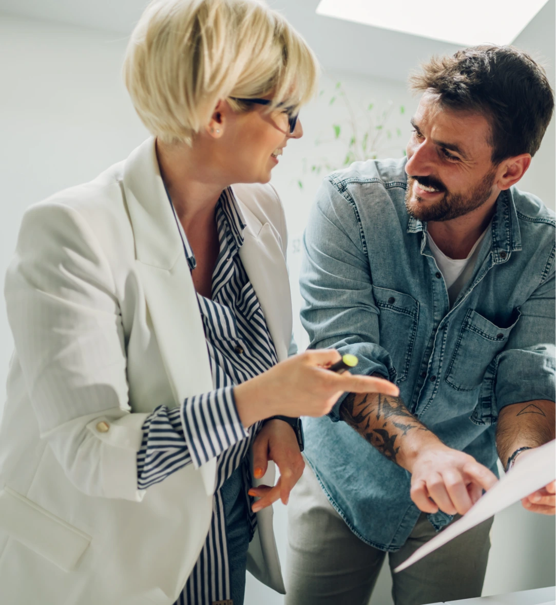 Zwei Menschen im Gespräch, eine Frau mit kurzem blondem Haar zeigt auf ein Dokument, ein Mann mit Bart und Tattoos lächelt.