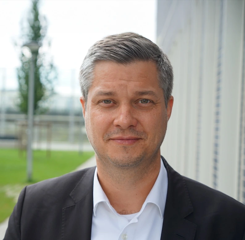 Porträt eines lächelnden Mannes mit kurzem grauem Haar, der einen schwarzen Anzug und ein weißes Hemd trägt, vor einem unscharfen Hintergrund mit Grünflächen und Gebäuden.