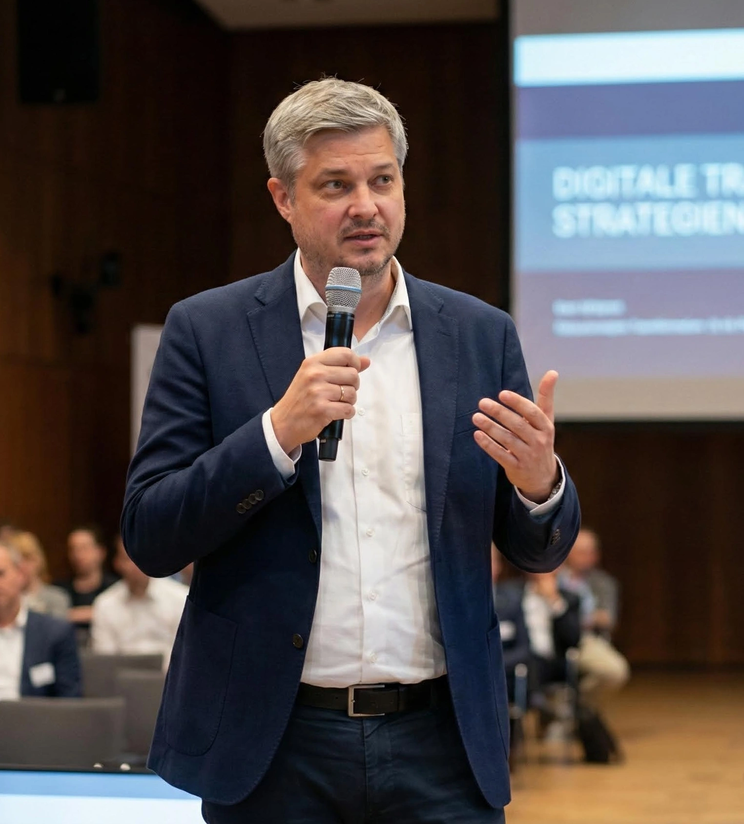 Mann in dunklem Anzug und weißem Hemd hält Mikrofon und spricht bei einem Vortrag mit verschwommenem Publikum im Hintergrund.