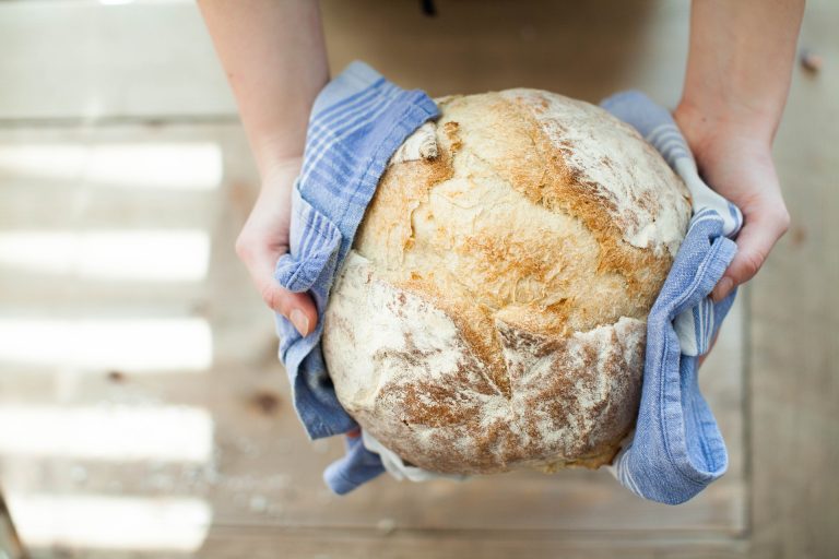 Frisch gebackenes rundes Brot, das mit einem blauen Küchentuch von zwei Händen gehalten wird.