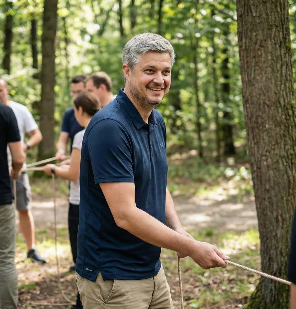 Lächelnder Mann in blauem Poloshirt hält ein Seil im Wald, andere Personen im Hintergrund.