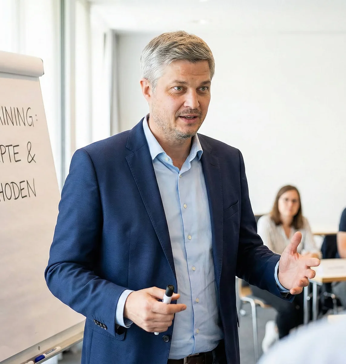 Ein Mann in dunkelblauem Anzug hält einen Marker und gibt in einem hellen Raum mit Flipchart eine Präsentation.