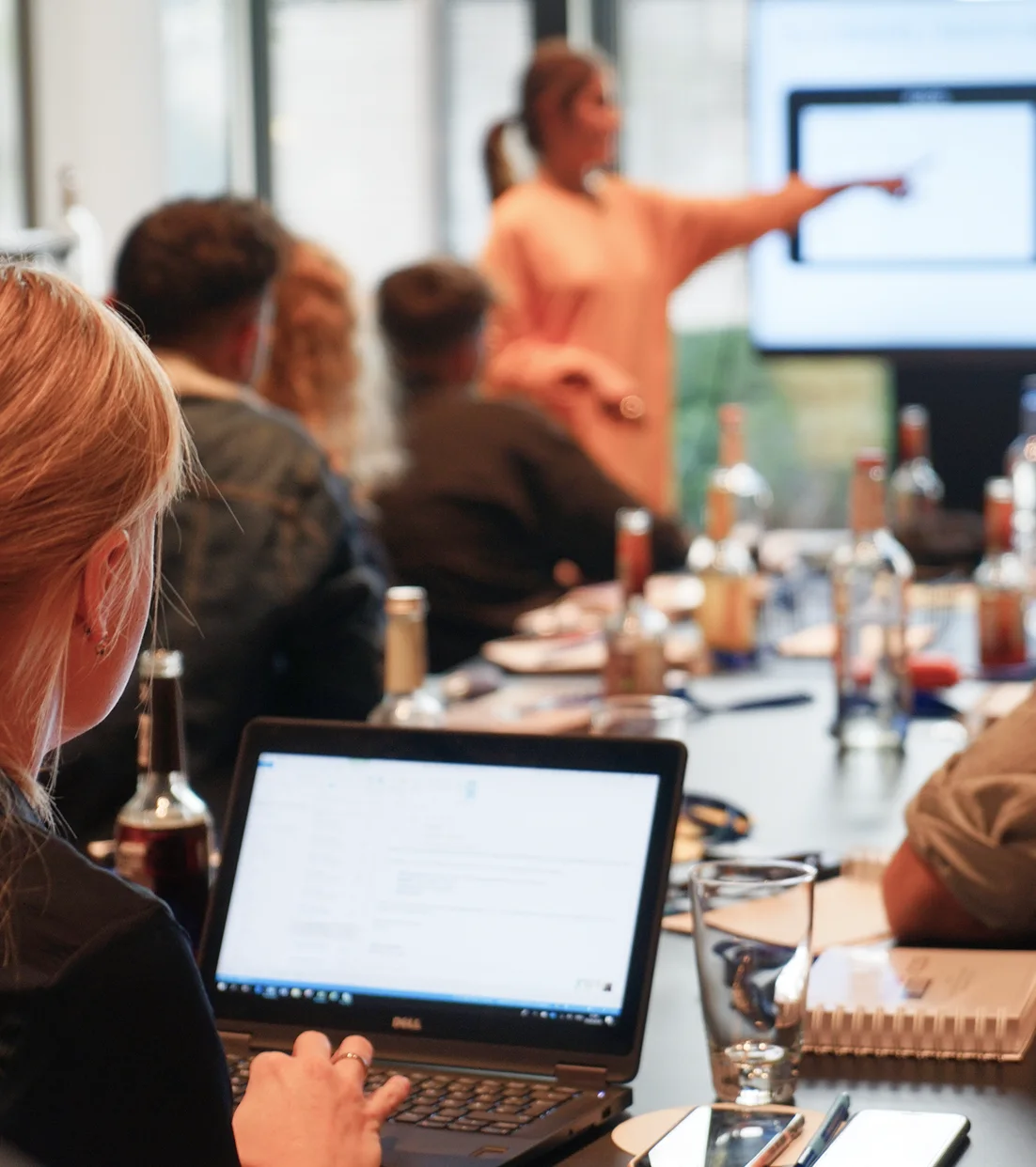 Frau arbeitet am Laptop in einem Meeting, während eine weitere Frau auf einen Bildschirm zeigt.
