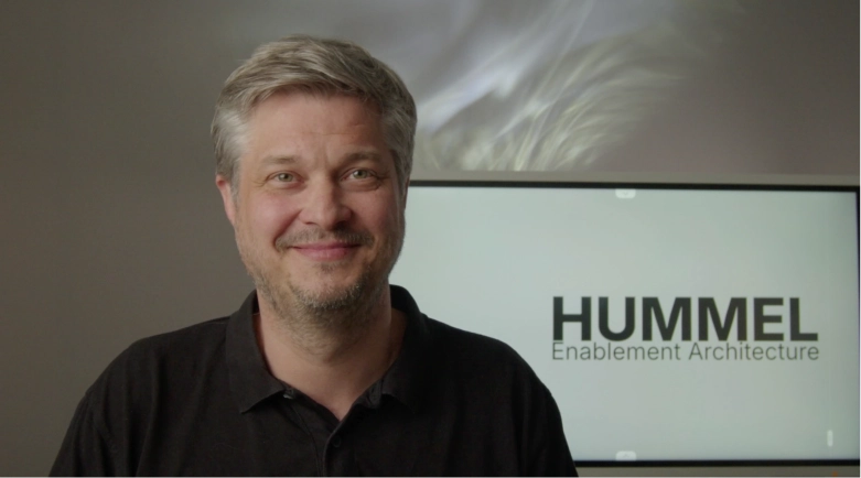 Ein lächelnder Mann mit grauem Haar vor einem Bildschirm mit der Aufschrift 'HUMMEL Enablement Architecture'.