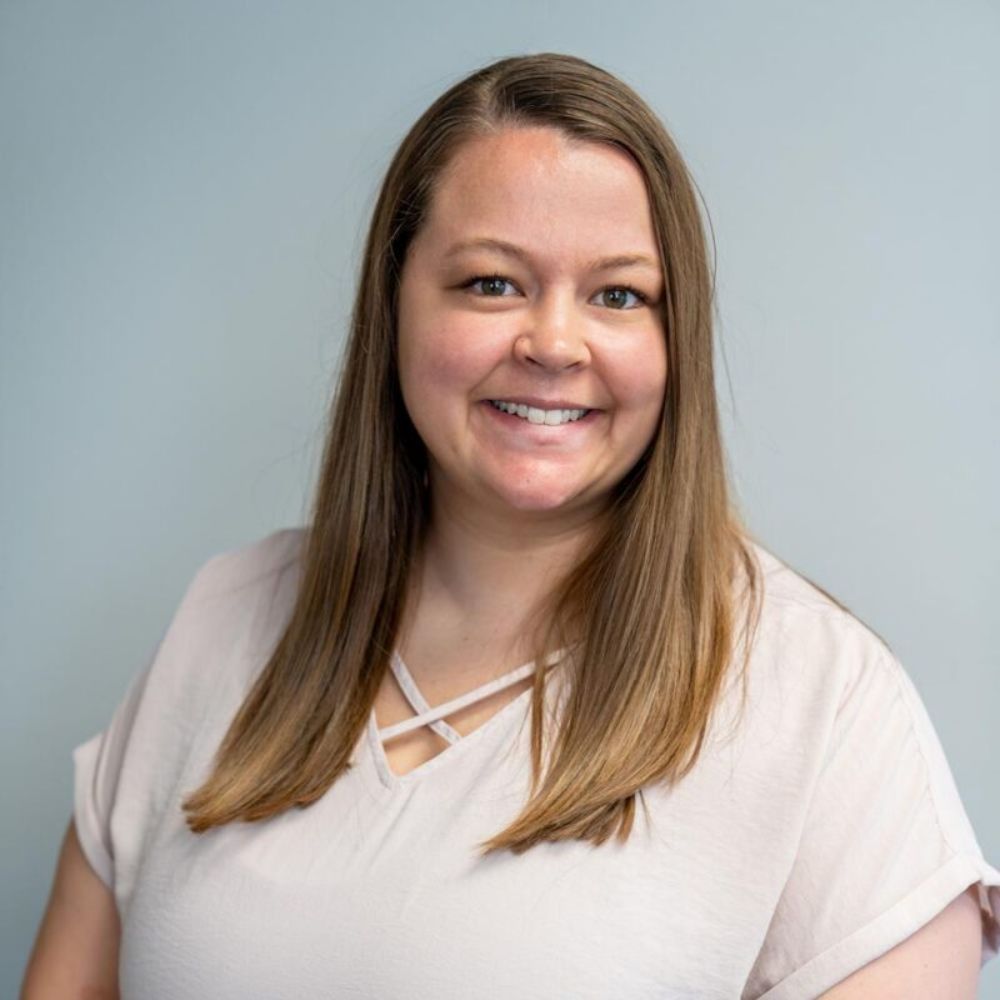 Smiling woman with long straight light brown hair wearing a light-colored top against a gray background.