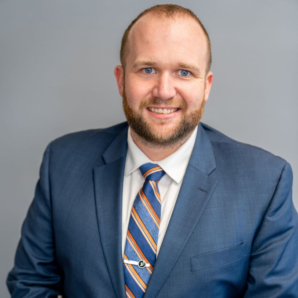 Smiling man with a beard wearing a blue suit, white shirt, and blue striped tie against a gray background.