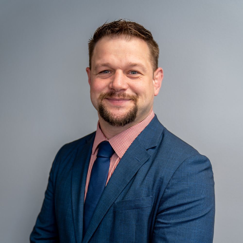 Smiling man with short brown hair and goatee wearing a blue suit, red checkered shirt, and navy blue tie against a gray background.