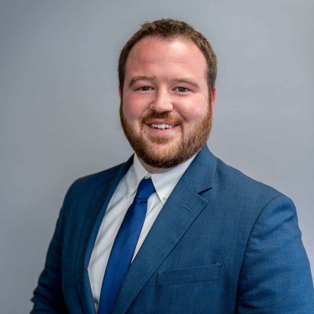Smiling man with beard wearing a blue suit jacket, white shirt, and blue tie against a gray background.