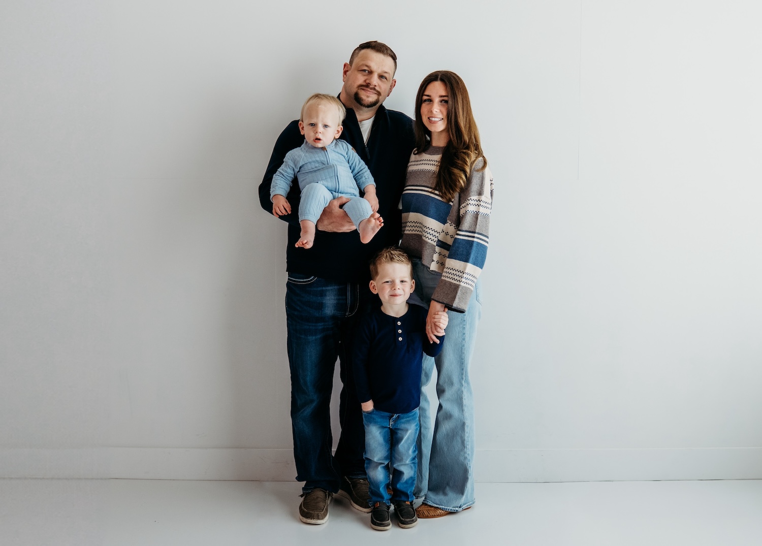 A family of four with a man holding a baby, a woman, and a young boy standing together against a plain white wall.