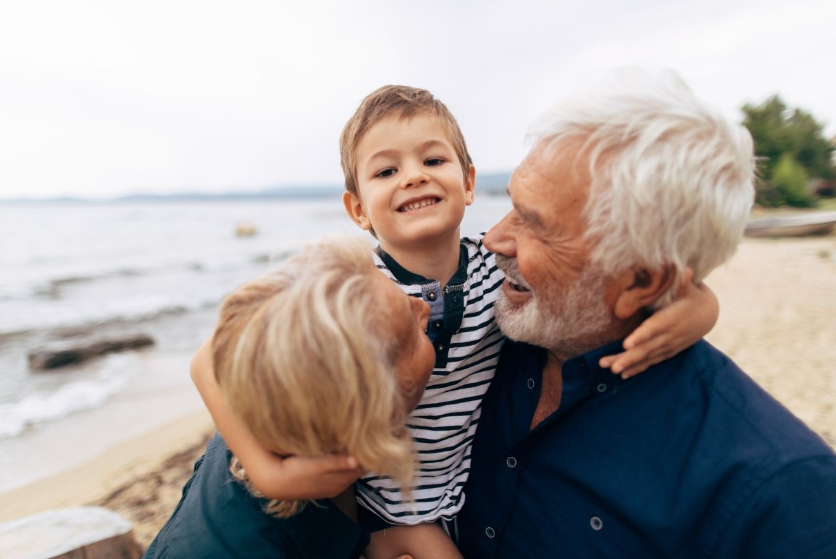 Smiling young boy being hugged by his grandparents on a beach.