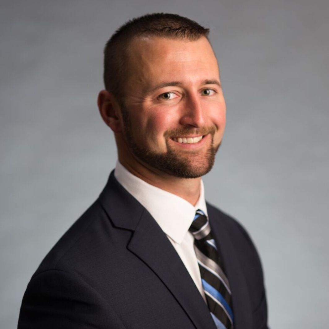 Smiling man with short hair and beard wearing a dark suit, white shirt, and striped tie against a gray background.