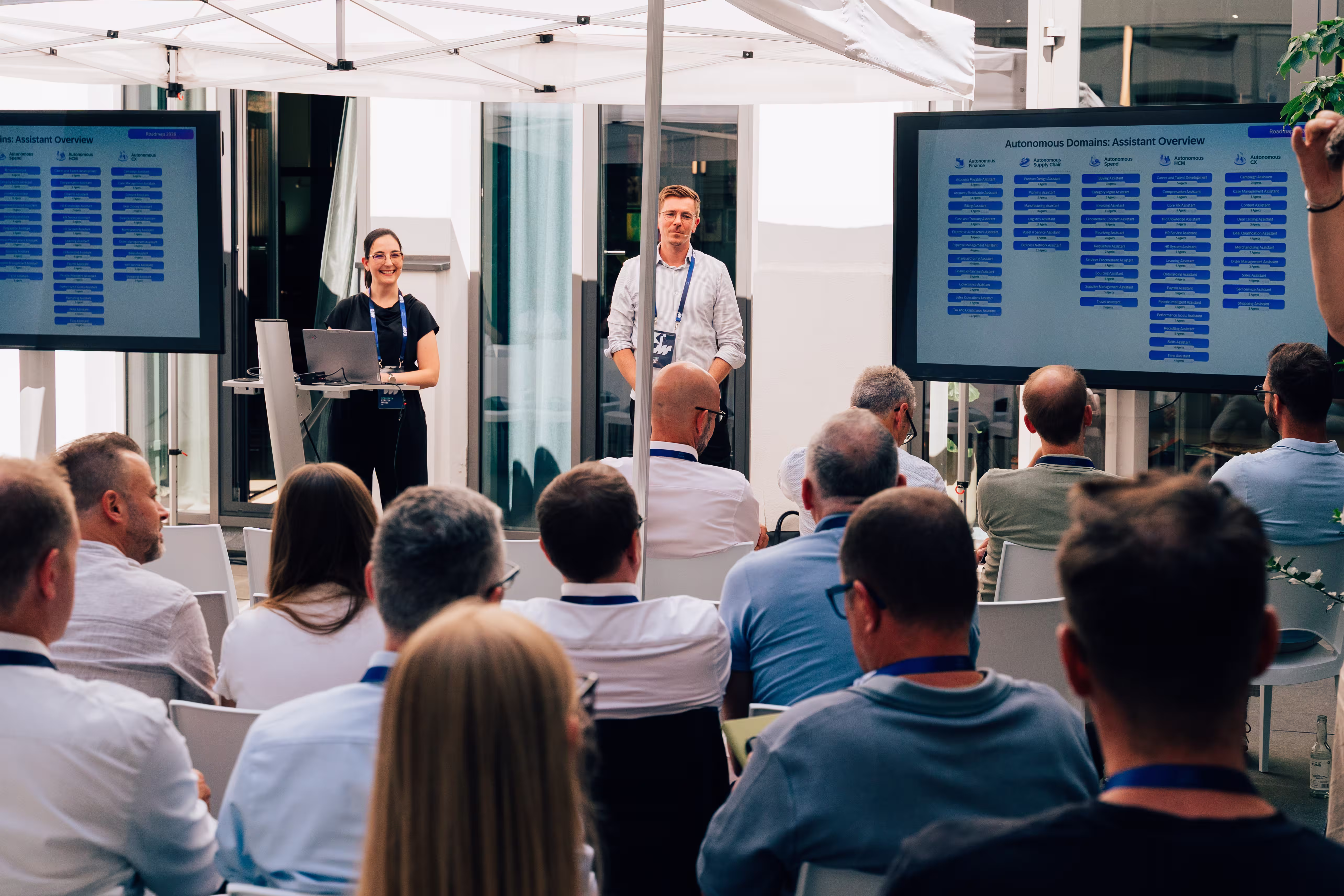 Two presenters introduce autonomous assistants to an audience, with an overview displayed on screens behind them.