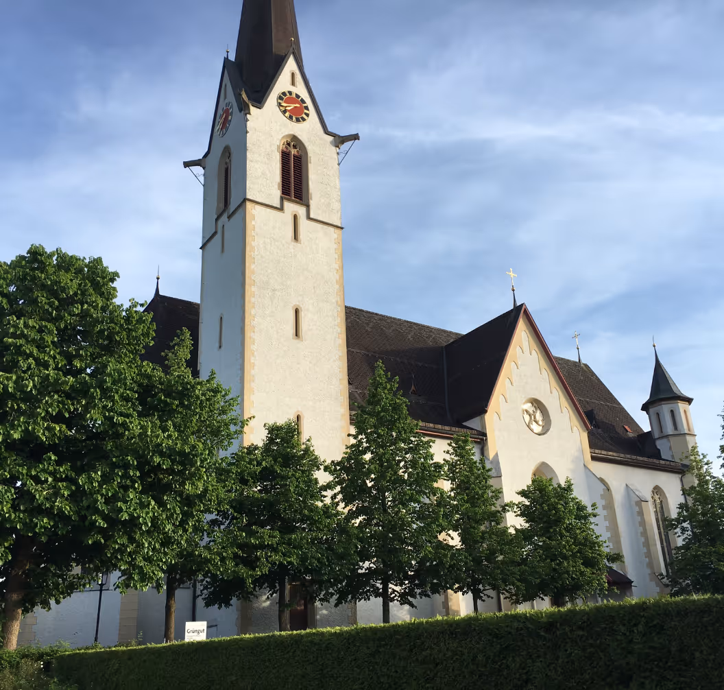 White church with a tall clock tower, steep roof, and surrounding green trees under a blue sky.