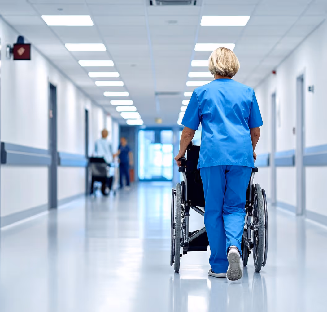 Healthcare worker in blue scrubs pushing an empty wheelchair down a hospital corridor.