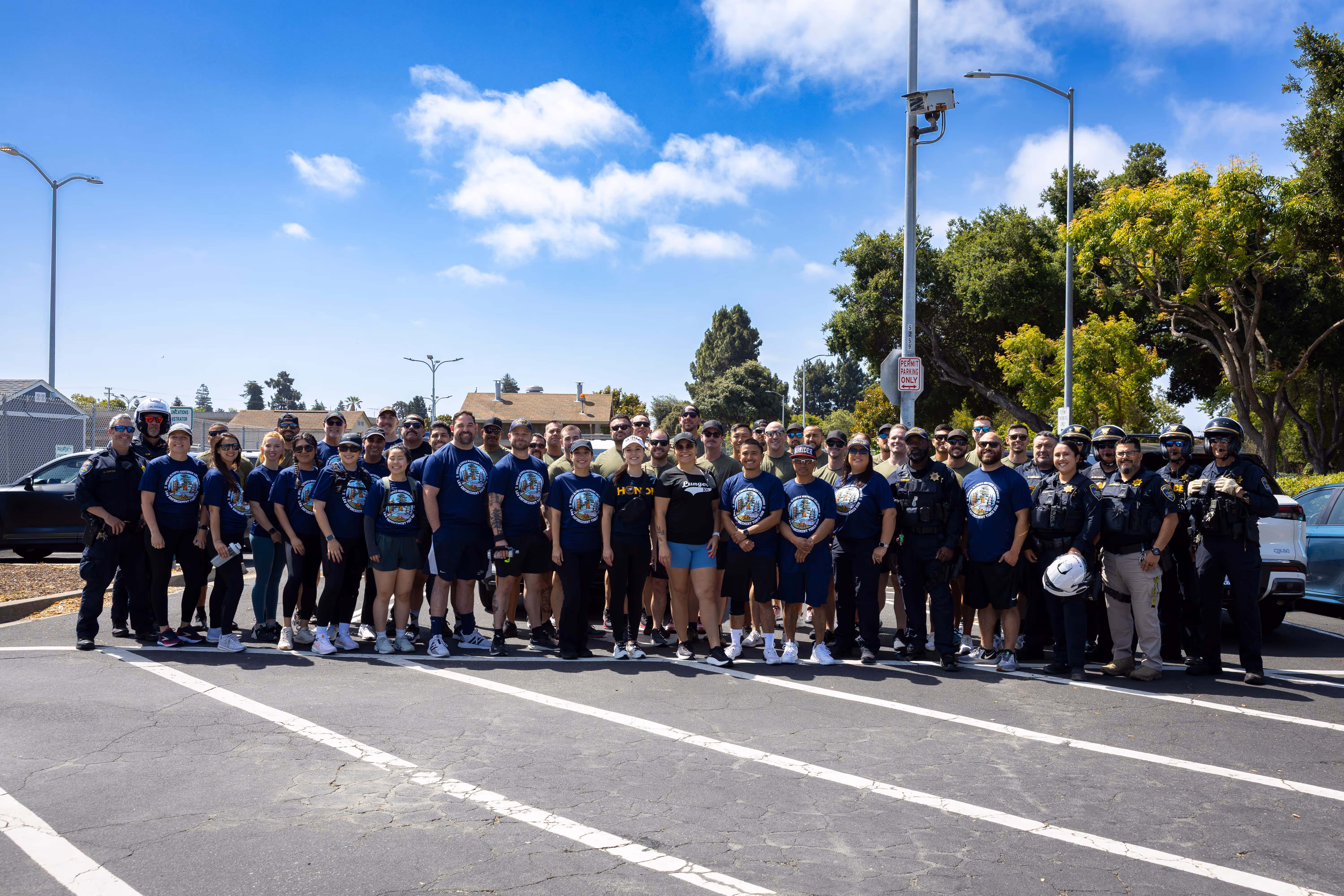 Large group of people, including police officers and civilians in blue event t-shirts, posing together outdoors on a sunny day.
