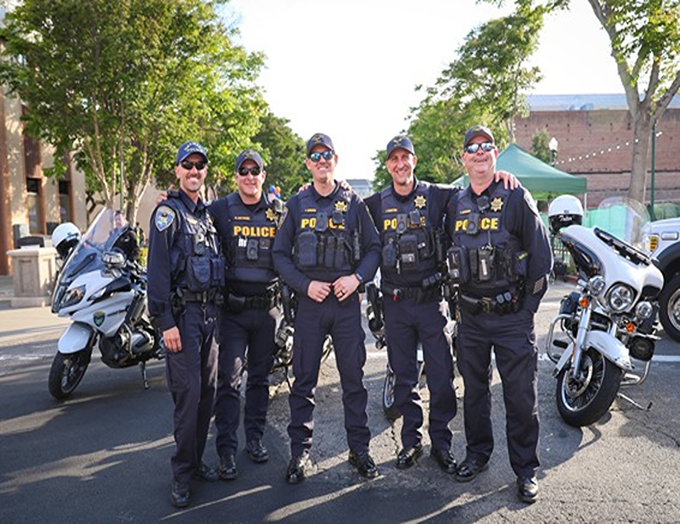 Five police officers in navy uniforms standing together smiling in front of two white police motorcycles on a sunny street.