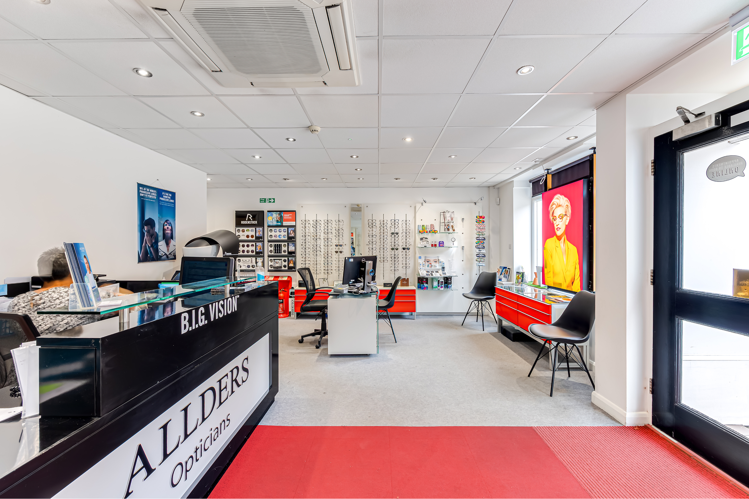 Modern optician store interior with "B.I.G. Vision" counter, eyeglass displays, and red accents.
