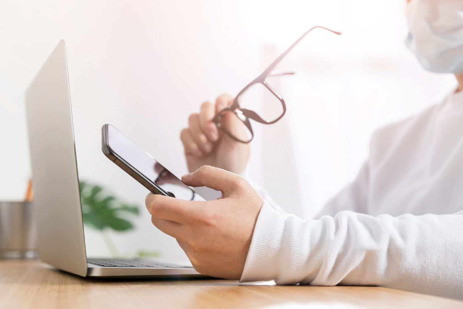 Person in white shirt holding phone and glasses near laptop at wooden desk.