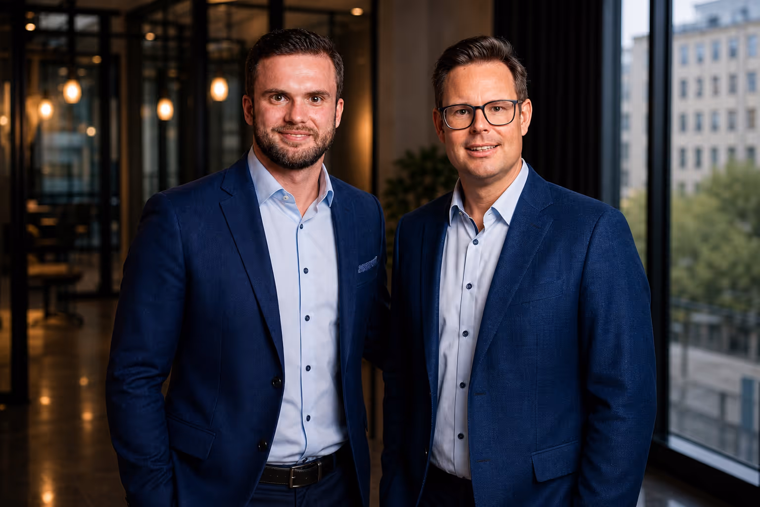 Zwei Männer im Büro tragen blaue Anzüge und Hemden, lächeln in die Kamera.