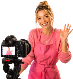 Junge Frau mit rosa Schürze winkt zur Kamera während der Aufnahme eines Videos.