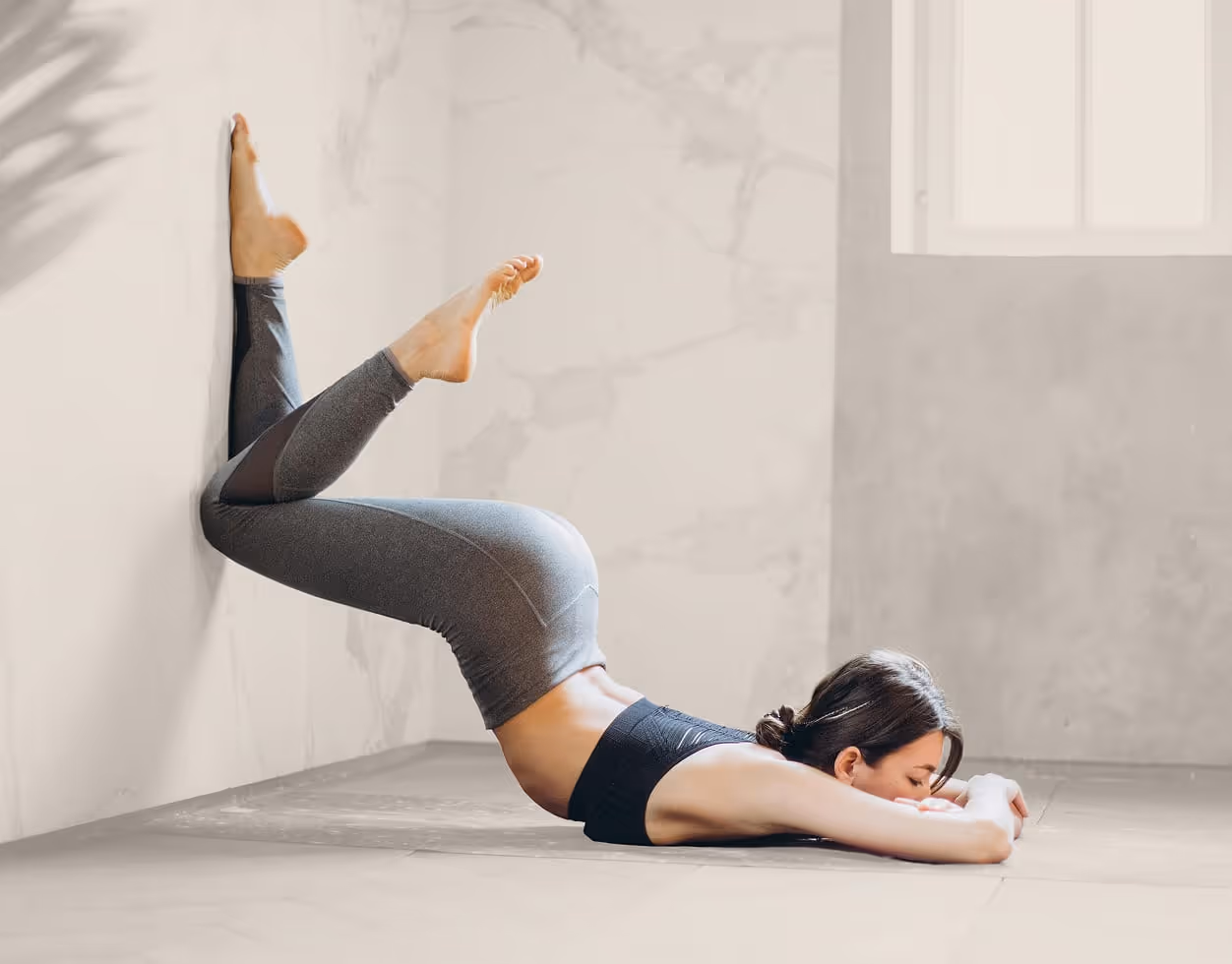 Woman lying face down on the floor with legs bent and feet pressed against a wall in a yoga stretch.