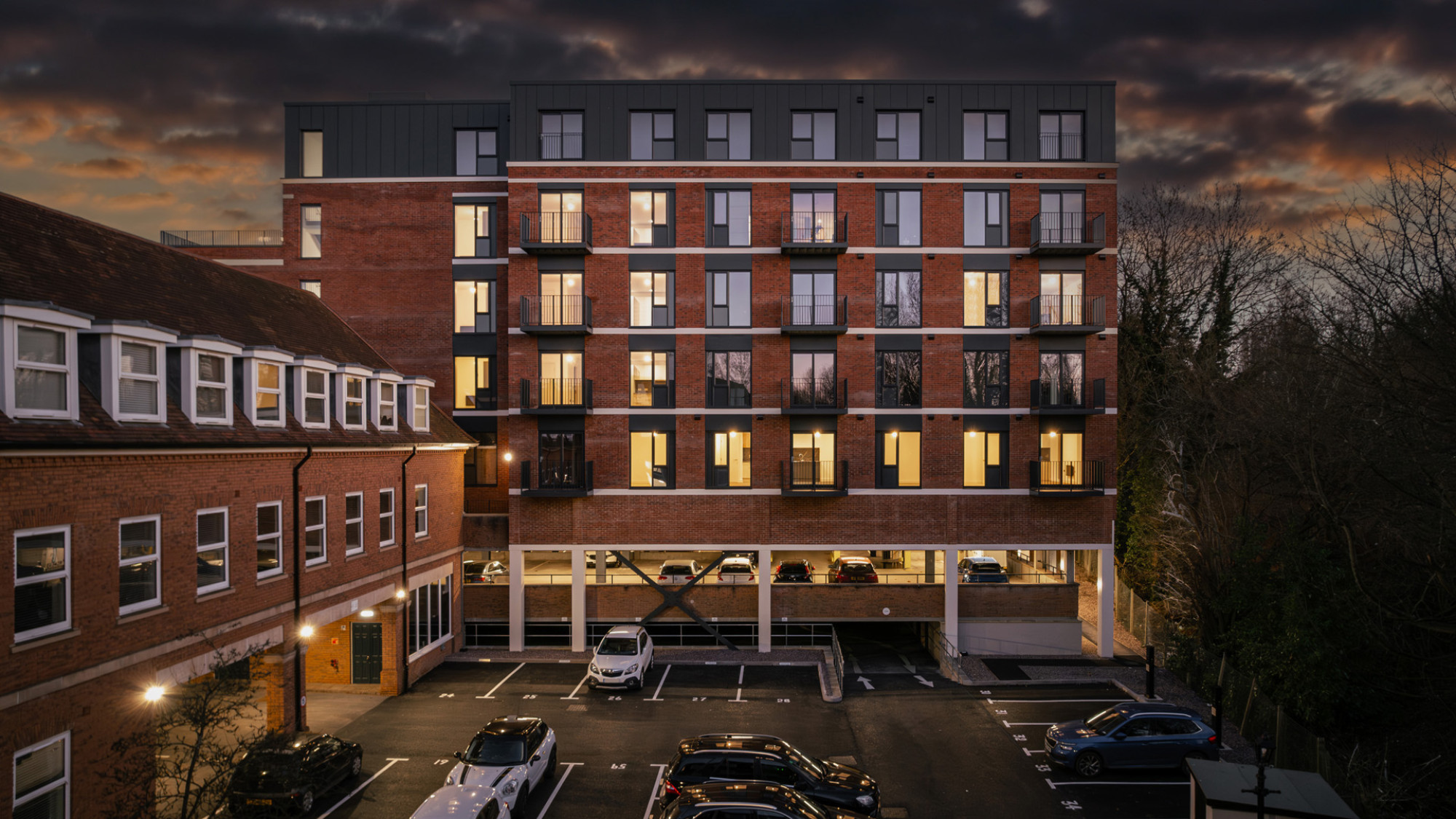 Apartment buildings by a calm river at sunset with colorful clouds and lights reflecting in the water.