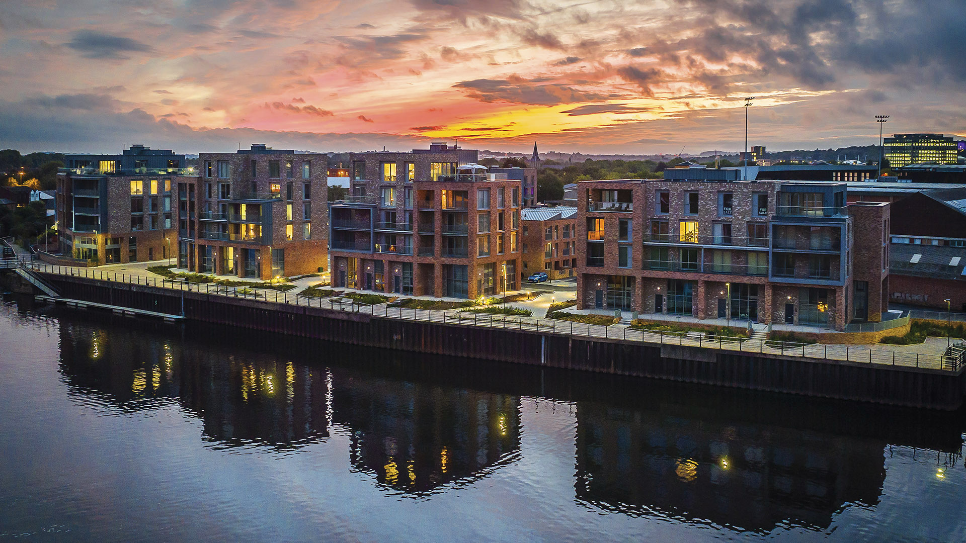 Apartment buildings by a calm river at sunset with colorful clouds and lights reflecting in the water.