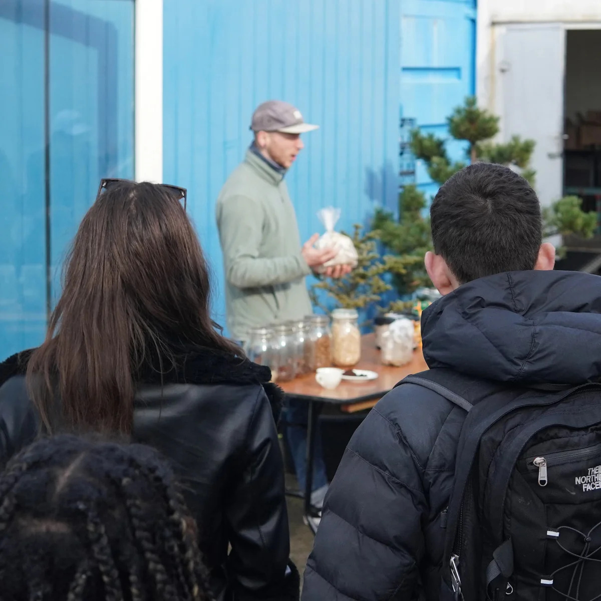 Man wearing a cap holding a bag, speaking to a small audience near a table with jars.