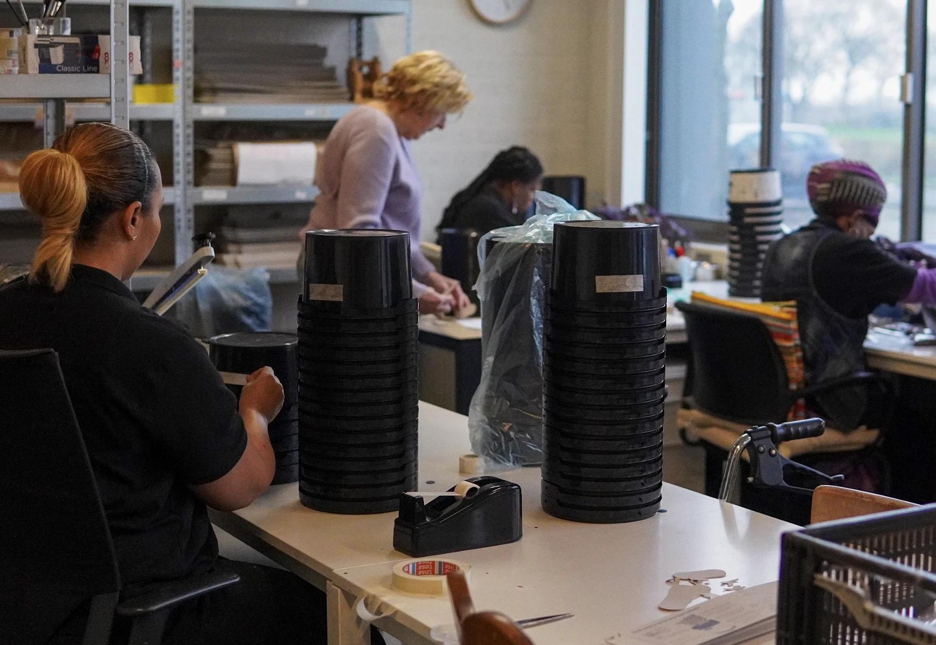 Four people working at tables organizing black plastic containers in a well-lit room with shelves and windows.